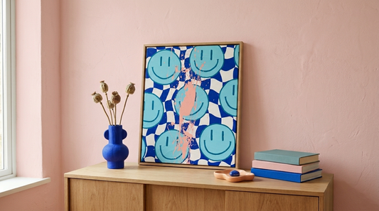 Farbenfrohe Dopamine Decor Wandkunst mit Smiley-Motiv in Blau und Pink, modern inszeniert auf einem Holzsideboard vor rosafarbener Wand, kombiniert mit Vase und Büchern im minimalistischen Interior