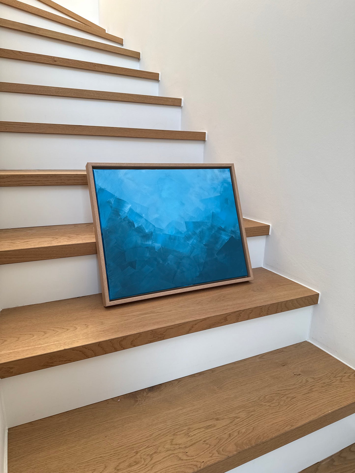 Abstraktes Acrylkunstwerk „BLUE“ in Blautönen, gerahmt in Naturholz, auf Holztreppe an heller Wand platziert.