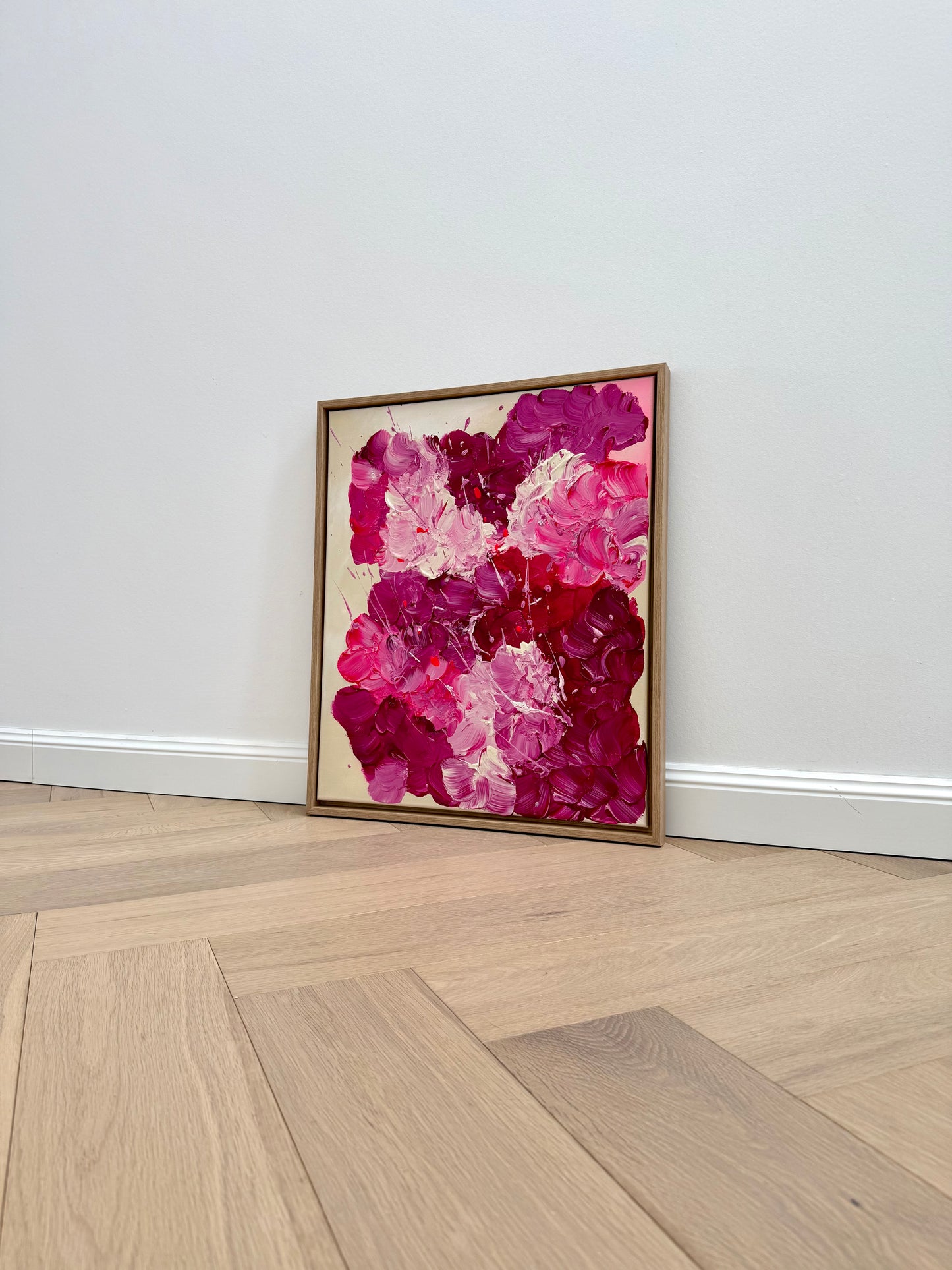 Abstraktes Blumenbild in 50x60 cm im Eiche-Rahmen, leicht schräg an eine helle Wand gestellt. Moderne florale Wandkunst mit kräftigen Pink- und Burgundertönen für stilvolle Wohnräume.