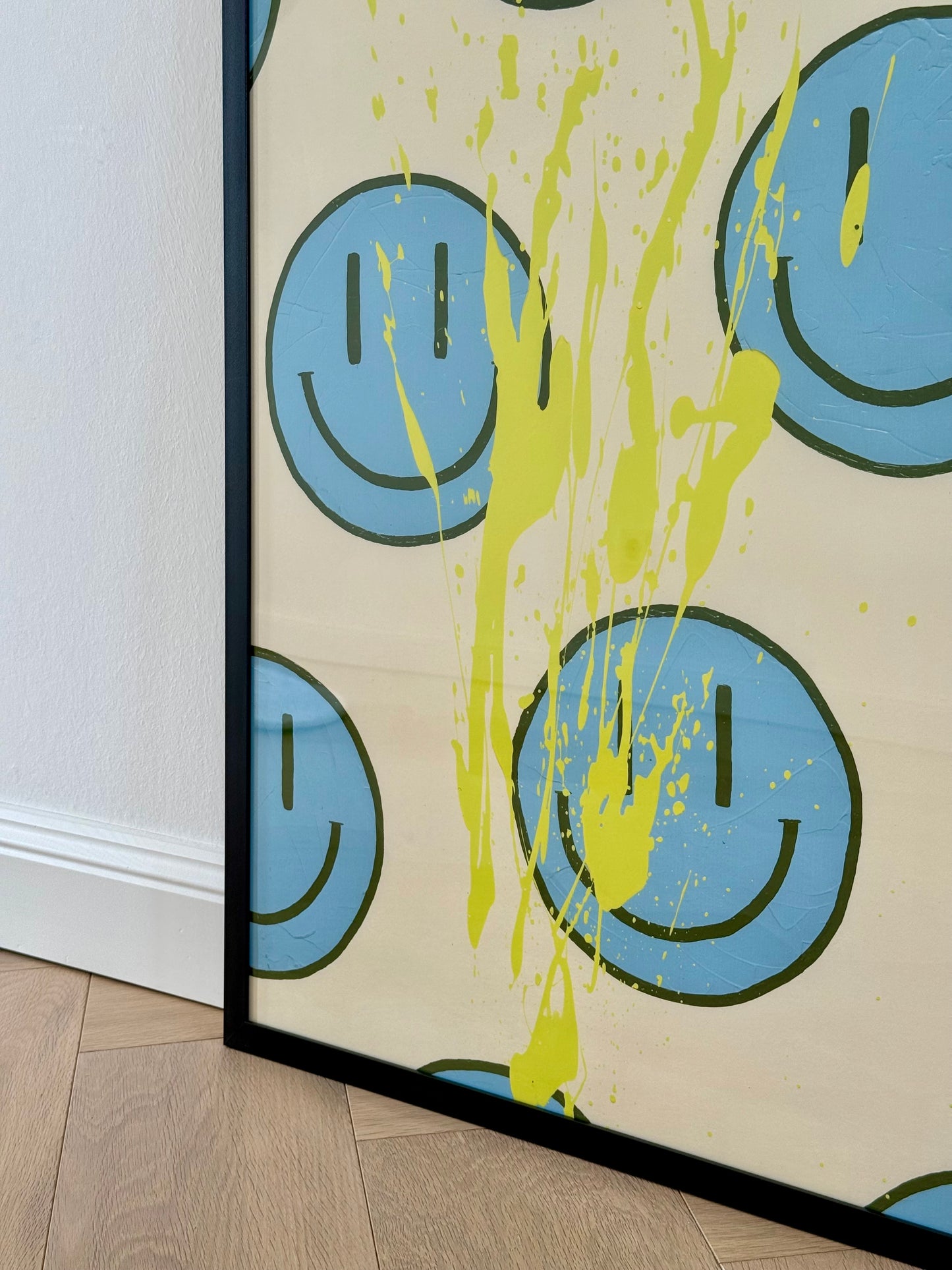 Seitliche Detailansicht des gerahmten Prints HAPPY #21 mit mehreren hellblauen Happy-Faces und leuchtend gelben Farbspritzern.