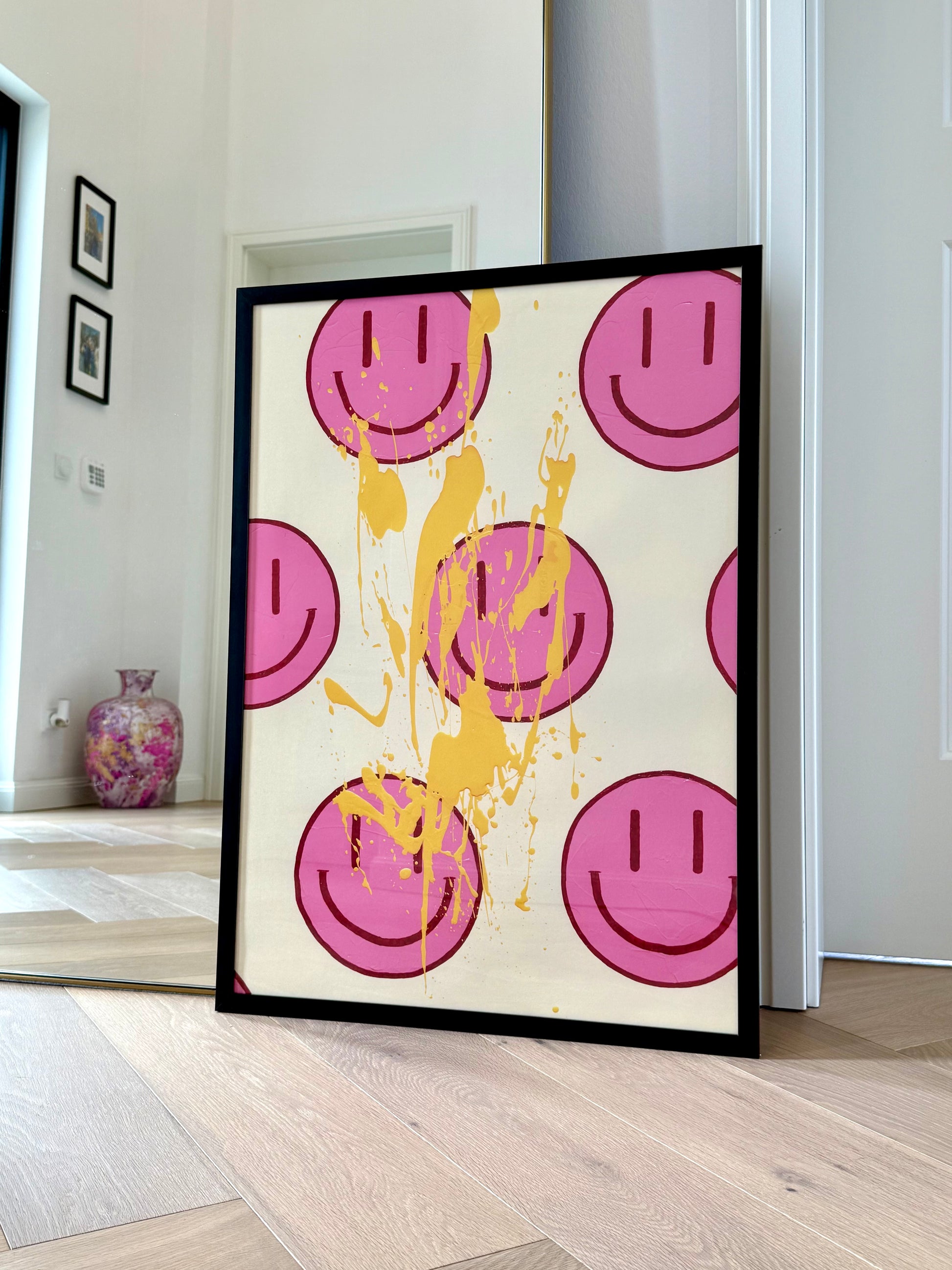 Der gerahmte Fine Art Print HAPPY #22 lehnt in einem hellen Wohnraum vor einem Spiegel und setzt mit Pink, Bordeaux und Hellorange einen kräftigen Farbakzent.