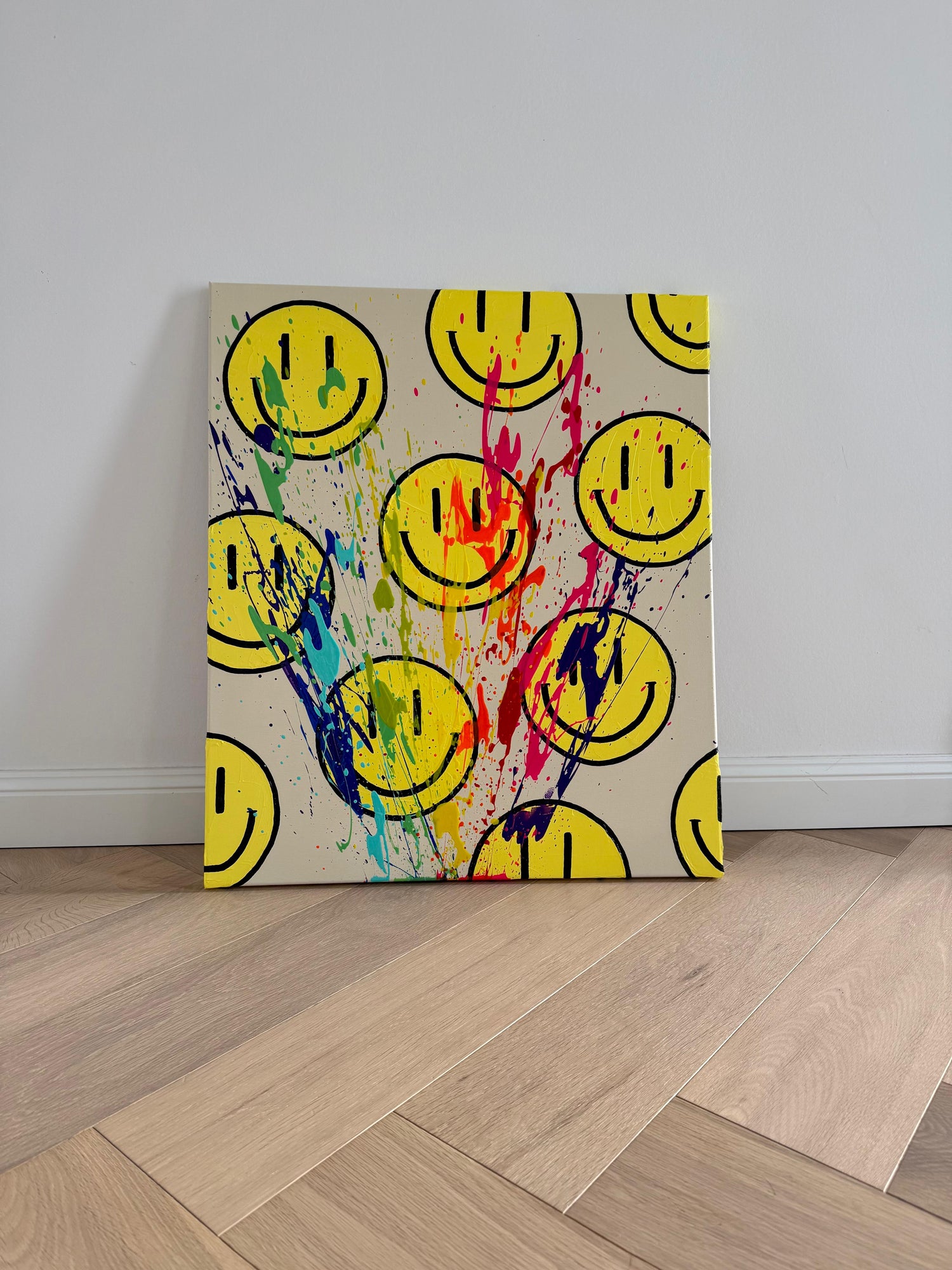 Abstraktes Acrylgemälde „HAPPY #11“ aus der HAPPY Series: gelbe Smiley-Motive auf hellem Hintergrund, überzogen von dynamischen Farbspuren in Pink, Blau, Grün und Violett. Das Kunstwerk lehnt ungerahmt an einer weißen Wand auf hellem Holzboden und verbindet grafische Elemente mit freier, expressiver Farbbewegung.