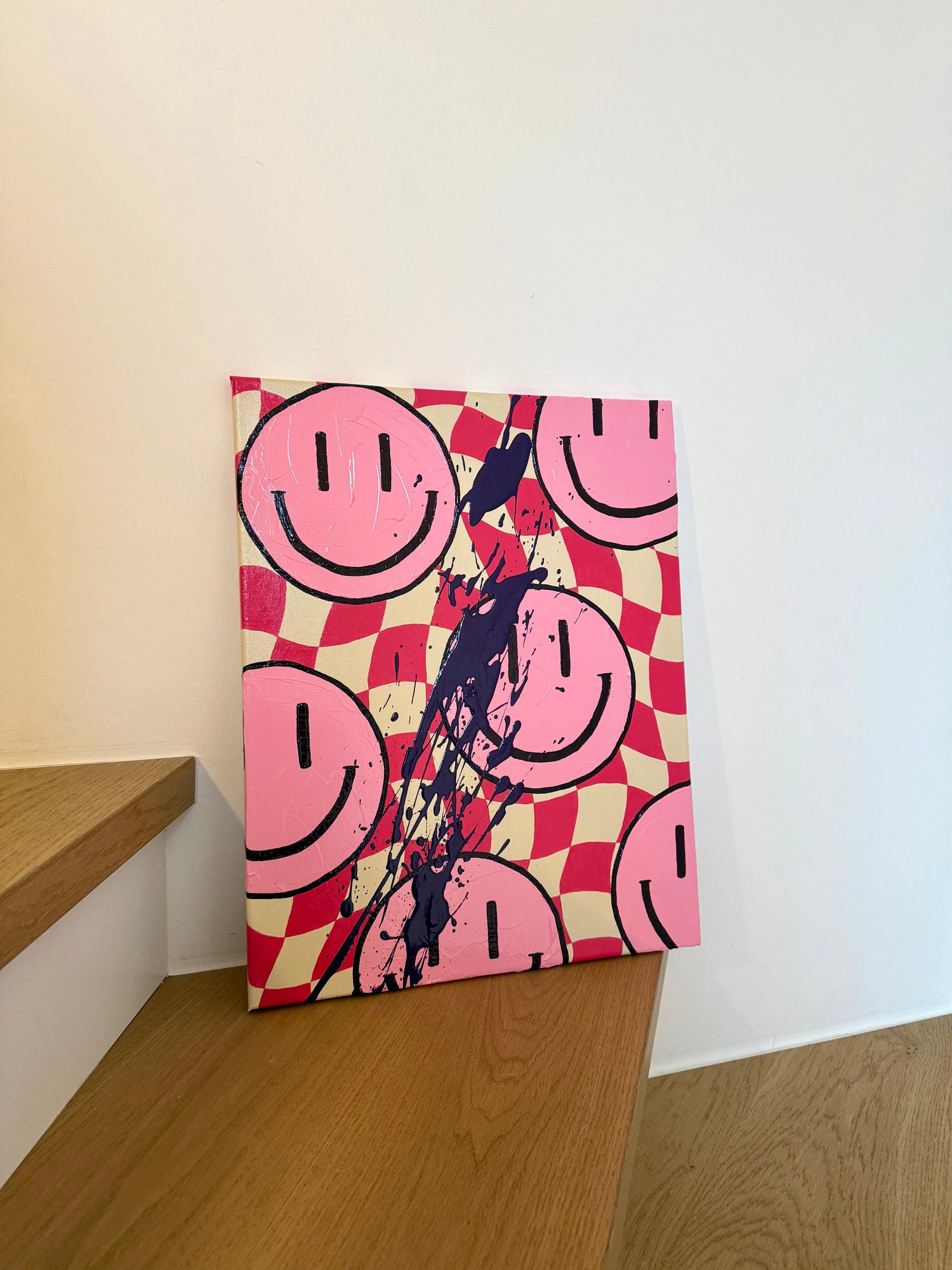 Abstraktes Acrylkunstwerk „HAPPY #12“ aus der HAPPY Series mit pinkem Schachbrett-Hintergrund, großflächigen Smiley-Motiven und expressiven Farbspritzern in Dunkellila und Pink. Das Werk lehnt auf einer Holztreppe und bringt starke Kontraste, Bewegung und energiegeladene Happy-Vibes in den Raum.