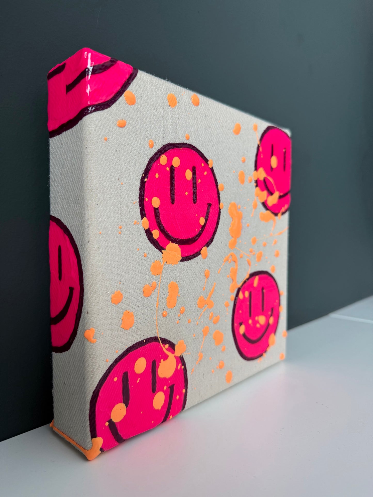 Seitliche Ansicht des Neonpink Smiley Kunstwerks „HAPPY #14“ mit apricotfarbenen Farbspritzern auf Naturleinwand