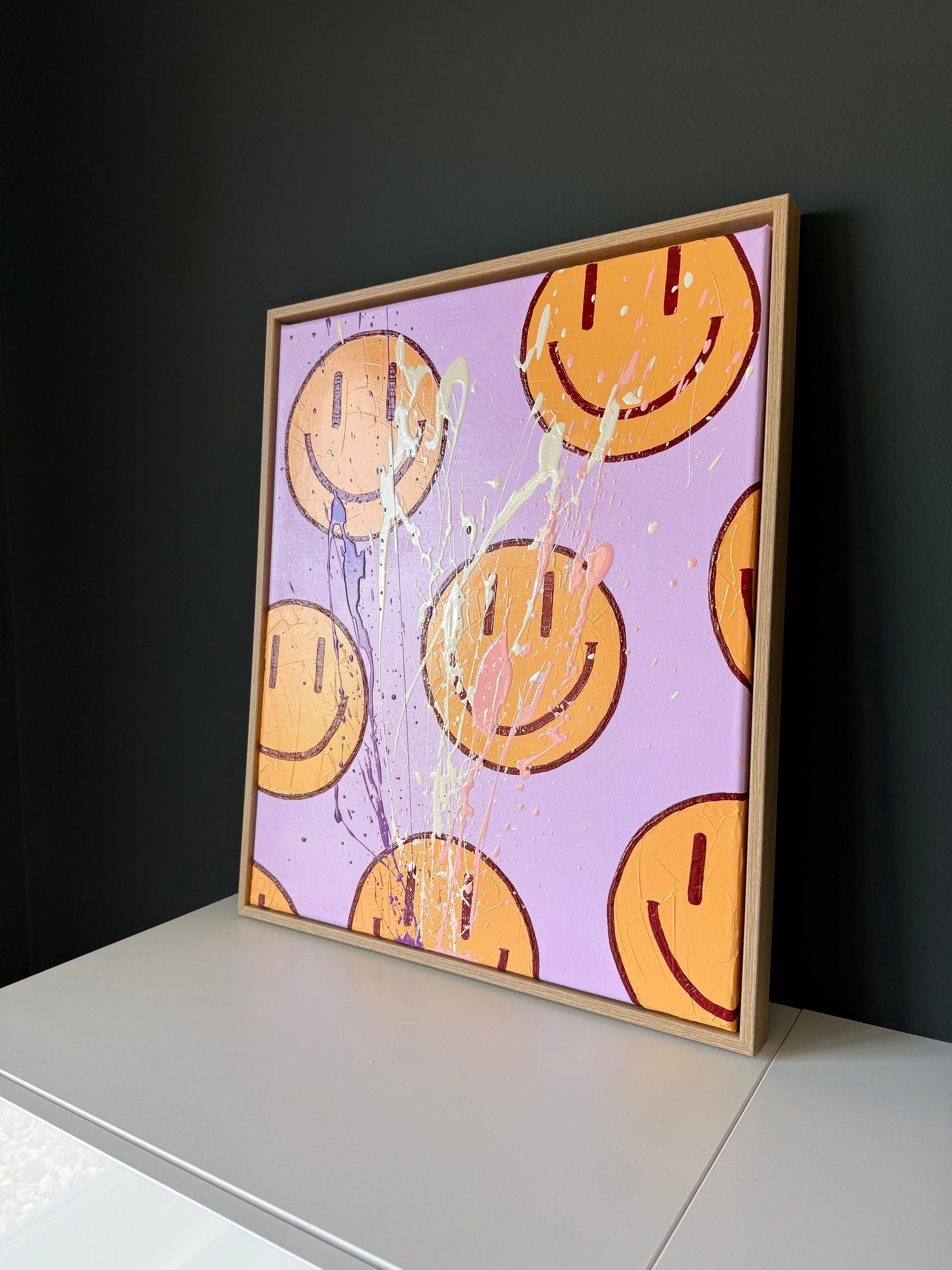 HAPPY #16, abstrakte Happy-Face Wandkunst in Flieder, Orange und Rosé mit Textur, Farbspritzern und glänzendem Finish im Raum inszeniert.