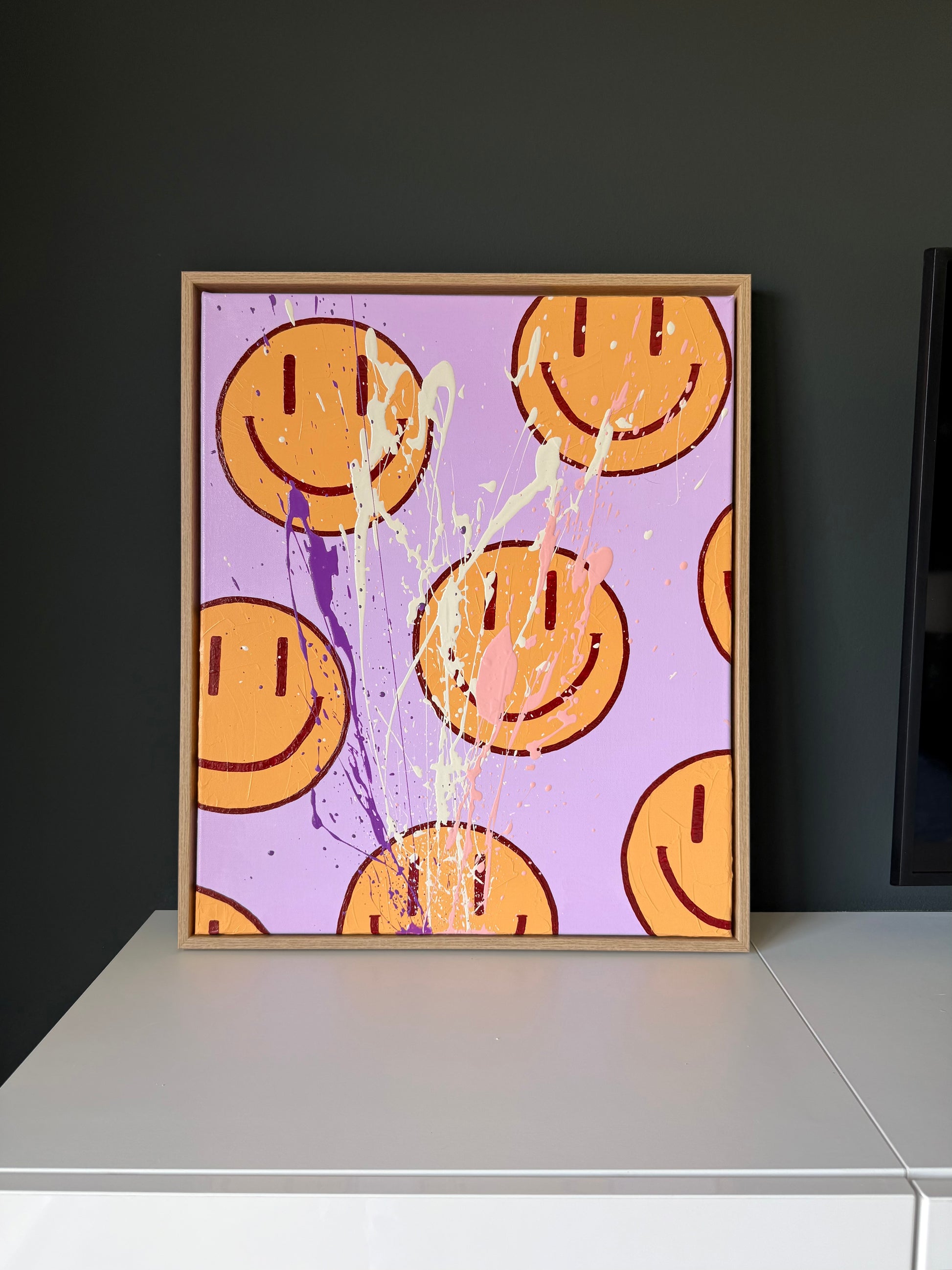 HAPPY #16, zeitgenössisches Happy-Face Kunstwerk auf Leinwand mit glänzender Oberfläche und Holzrahmen vor dunkler Wand.