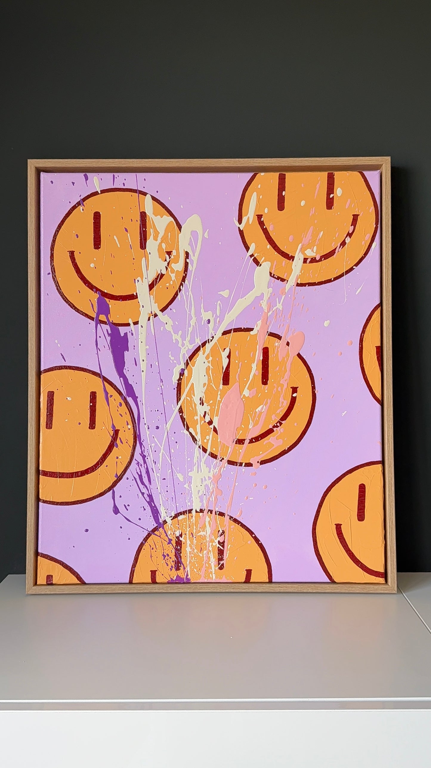 HAPPY #16, abstrakte Happy Face Wandkunst in Flieder, Orange und Rosé mit Textur, Farbspritzern und glänzendem Finish auf einem Sideboard positioniert.