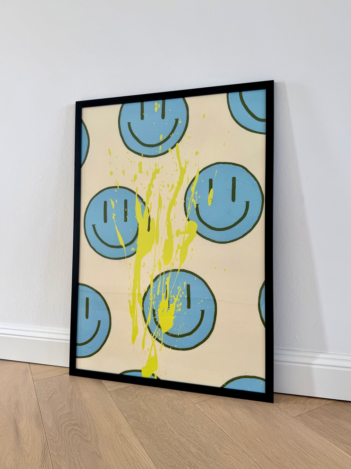 Gerahmter Fine Art Print HAPPY #21 lehnt an einer hellen Wand auf Holzboden. Hellblaue Happy-Faces und zitronengelber Farbspritzer treffen auf einen cremefarbenen Hintergrund.