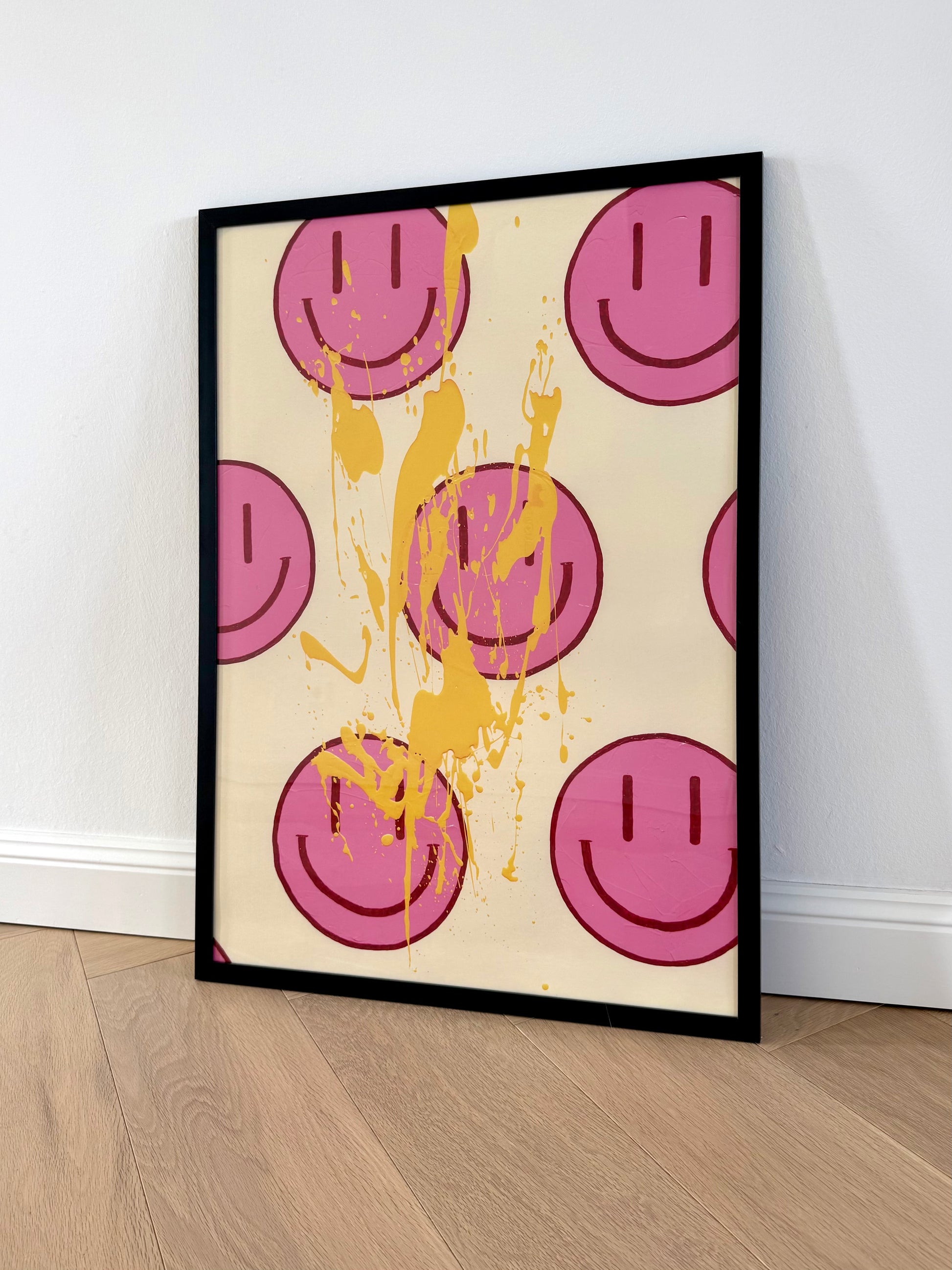 Gerahmter Fine Art Print HAPPY #22 lehnt an einer hellen Wand auf Holzboden. Pinkfarbene Happy-Faces und hellorange Farbspritzer treffen auf einen cremefarbenen Hintergrund.
