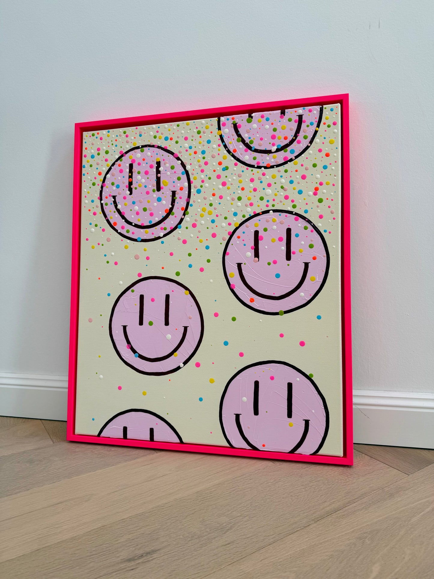 Schräg fotografiertes Originalkunstwerk HAPPY #26 mit hellrosa Happy Faces, bunten Konfettipunkten und leuchtendem neonpinkem Rahmen.