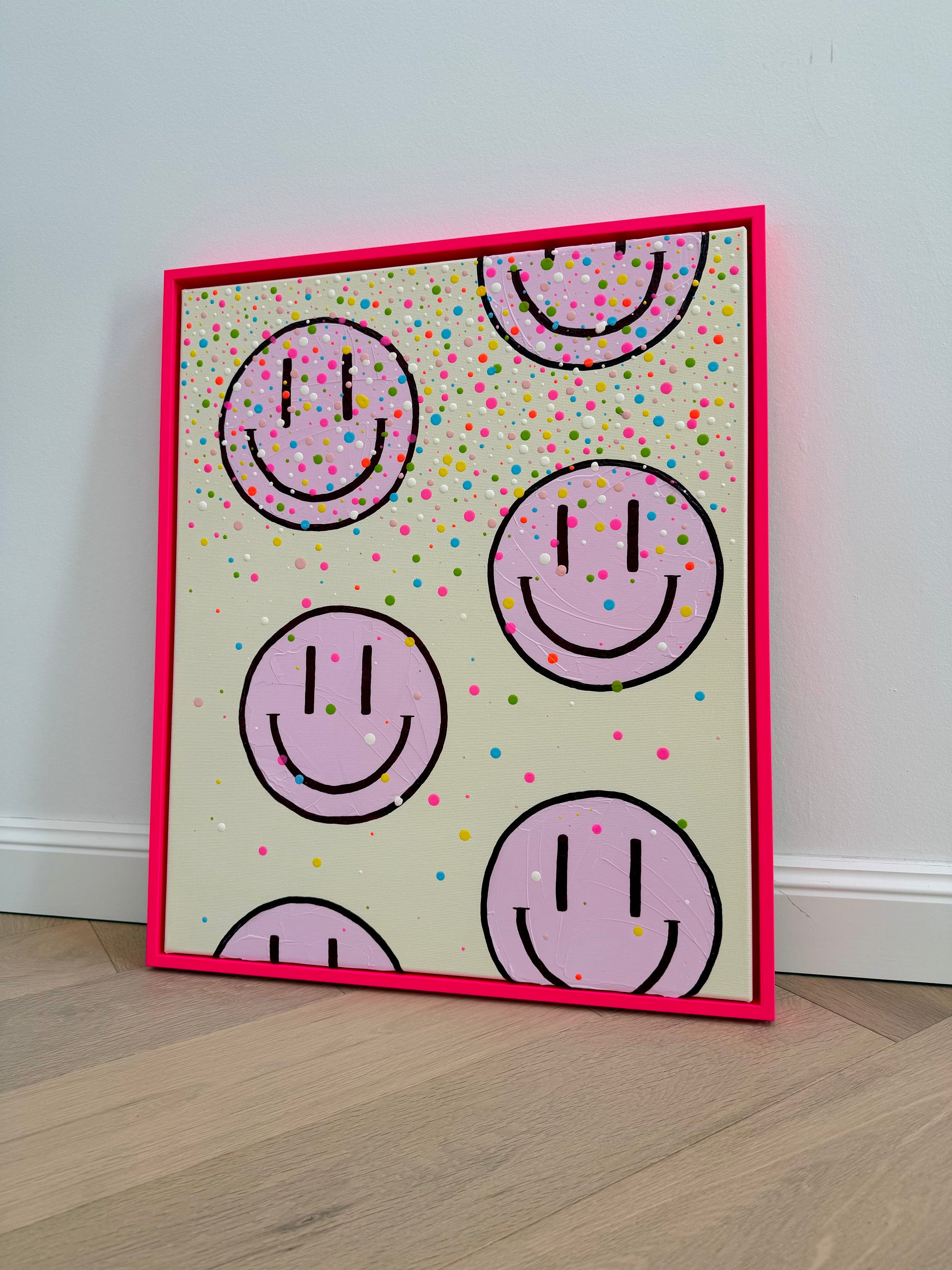 Schräg fotografiertes Originalkunstwerk HAPPY #26 mit hellrosa Happy Faces, bunten Konfettipunkten und leuchtendem neonpinkem Rahmen.