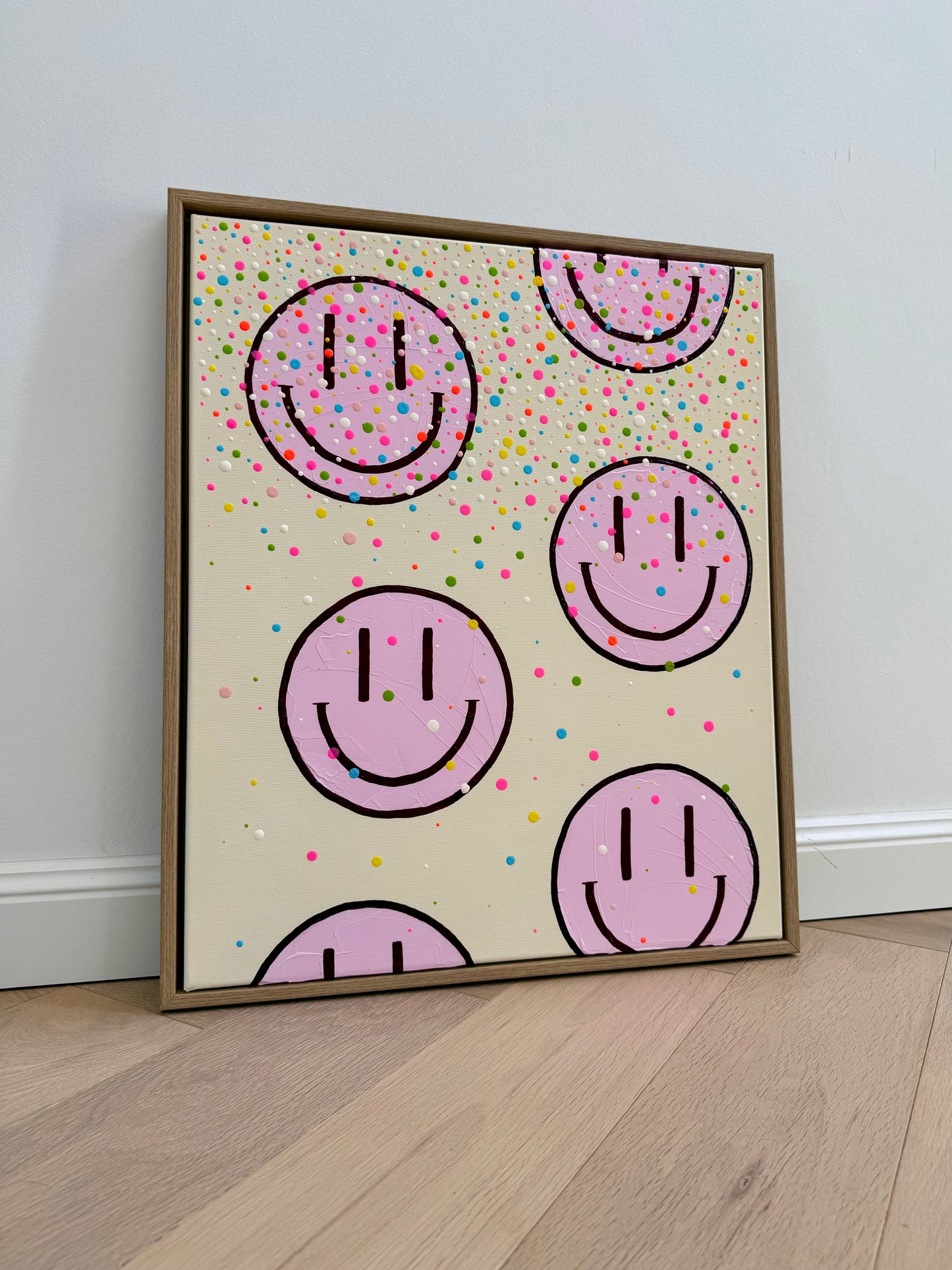 Handgemaltes Acrylbild HAPPY #26 mit hellrosa Happy Faces und buntem Konfetti, schräg fotografiert im Schattenfugenrahmen aus Eiche Natur.