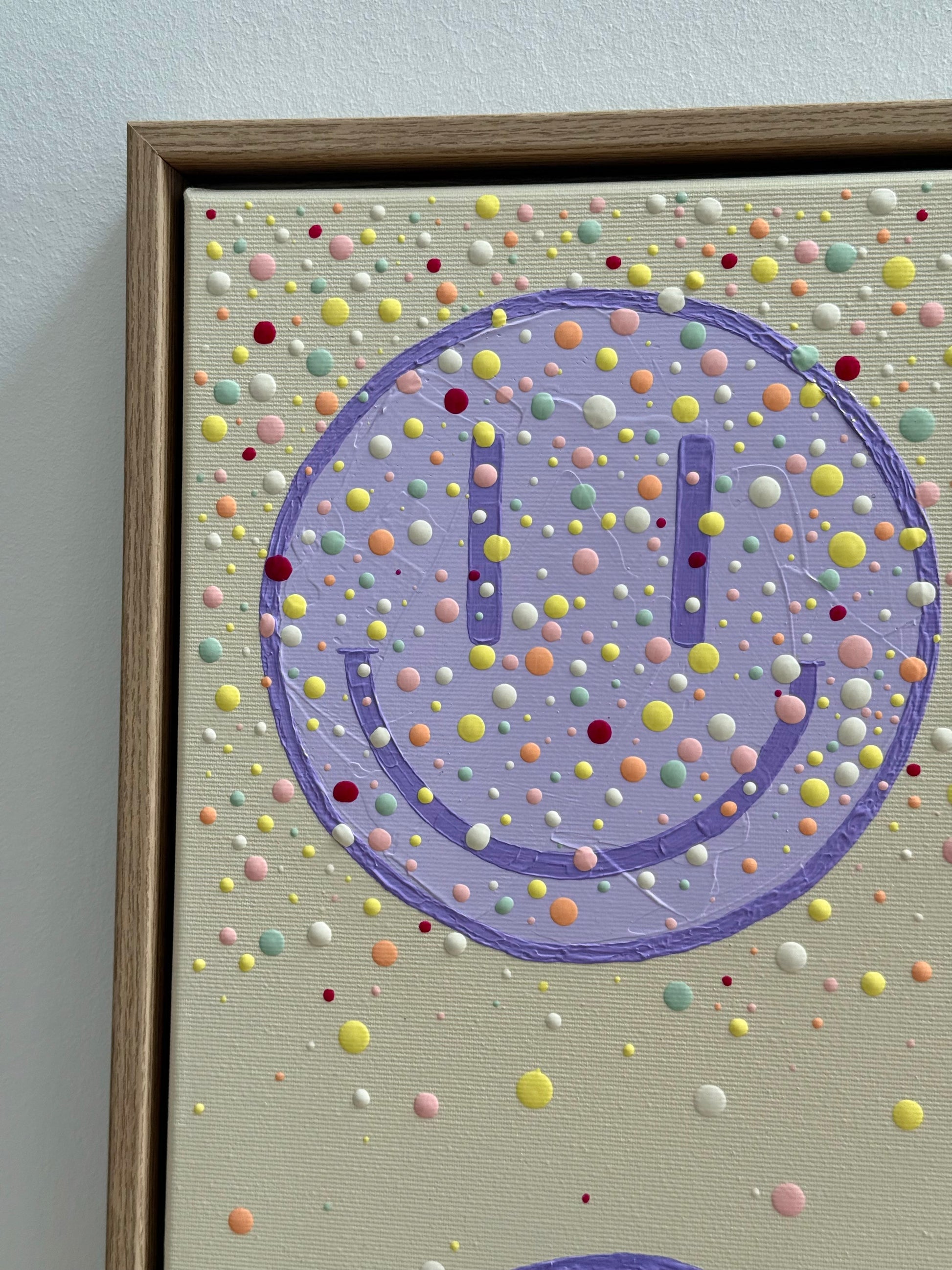 Nahaufnahme eines fliederfarbenen Happy-Faces mit sichtbarer Acrylstruktur und handgesetzten Farbpunkten auf beigem Hintergrund.