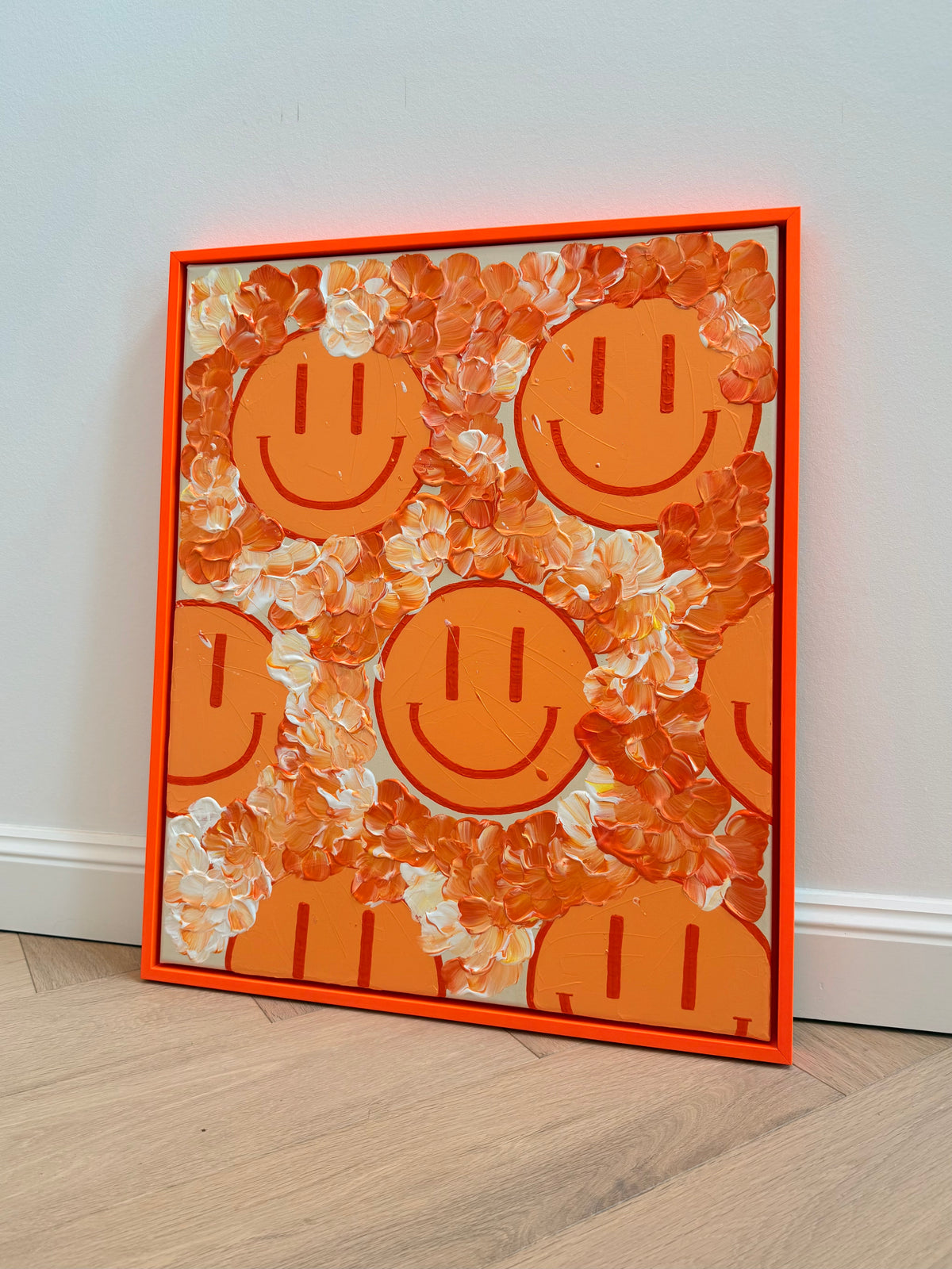 Schrägansicht von HAPPY #30 mit pastellorangefarbenen Happy Faces, plastischer Blütenstruktur und leuchtendem orangefarbenem Rahmen, angelehnt an eine helle Wand.