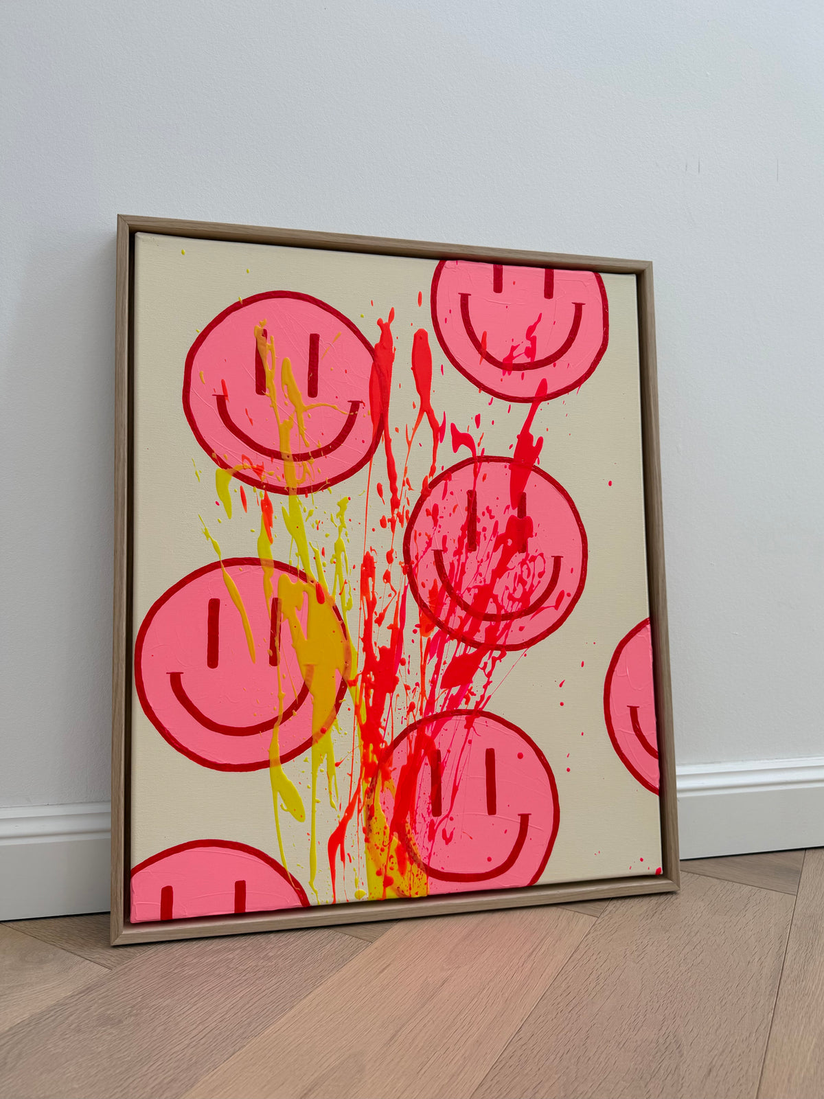 HAPPY #32 mit pinken Happy Faces, kirschroten Konturen und Farbspritzern in Zitronengelb, Neonorange und Neonpink auf beigem Grund, gerahmt in Eiche Natur.