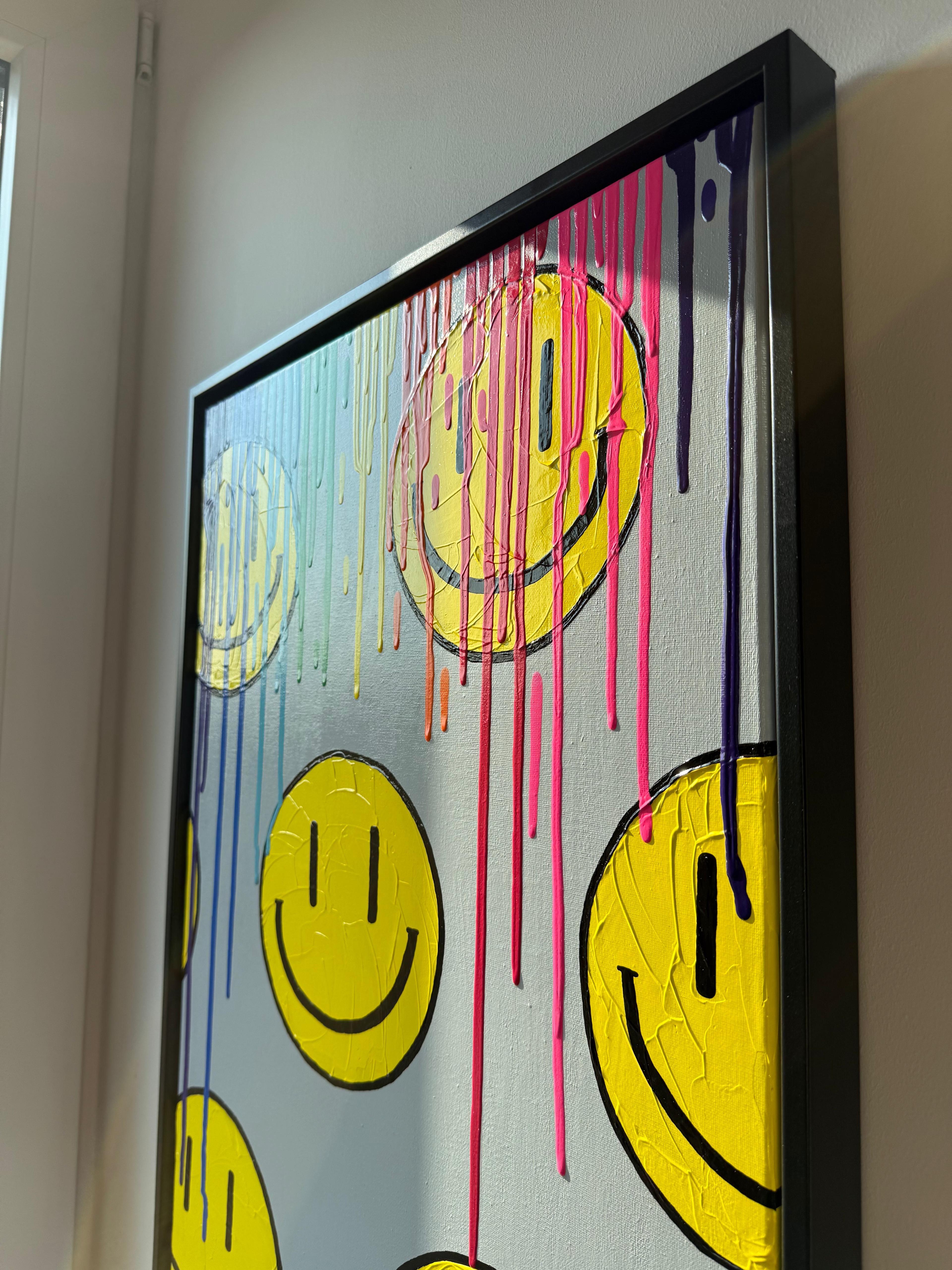 Seitliche Ansicht eines abstrakten Smiley-Kunstwerks mit leuchtend gelben Gesichtern und farbigen Farbverläufen in Regenbogenfarben. Das gerahmte Bild wirkt lebendig, verspielt und setzt einen fröhlichen Farbakzent an der Wand.