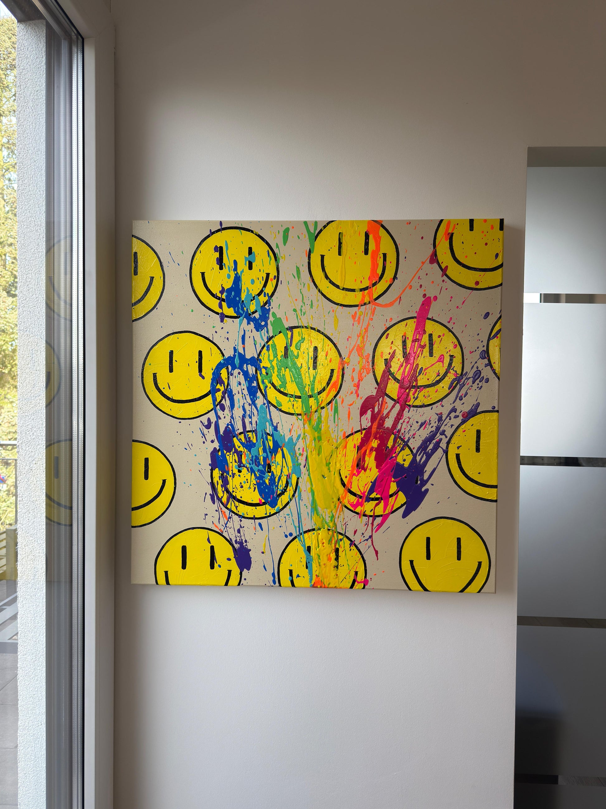 Abstraktes Acrylkunstwerk HAPPY #7 aus der HAPPY Series von Sophia Toschka, 85×85 cm, Acryl auf Leinwand. Gelbe Smiley-Motive auf hellem Hintergrund, kombiniert mit dynamischen Farbspritzern in Blau, Pink, Grün und Gelb, als großformatiges Wandkunstwerk in einem modernen, hellen Raum inszeniert.