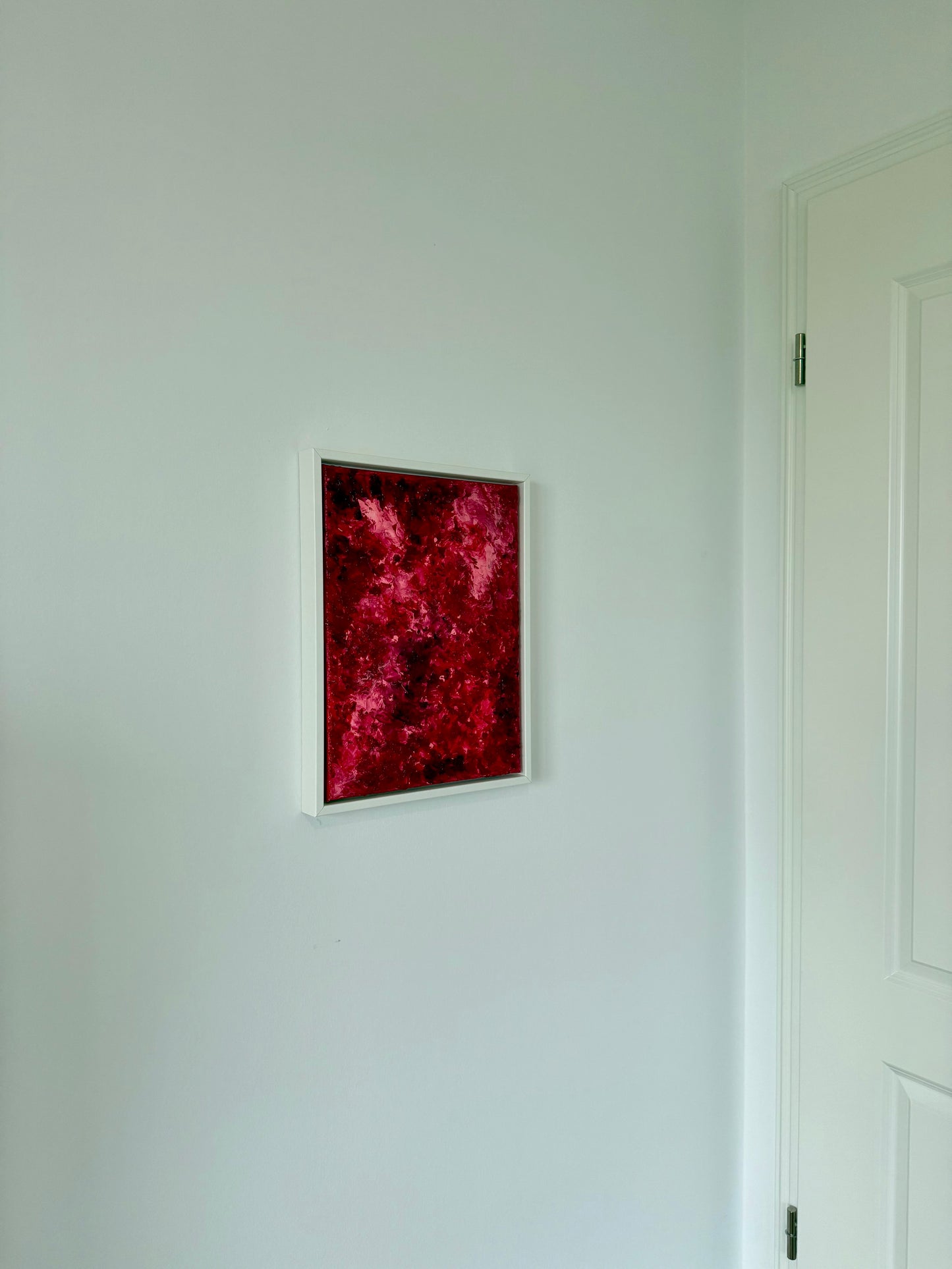 Kleines abstraktes Kunstwerk in kräftigen Rot- und Pinktönen, gerahmt in Weiß, an einer hellen Wand im Wohnraum aufgehängt. Das Bild setzt einen intensiven Farbakzent in einem minimalistischen, ruhigen Interior und wirkt modern sowie klar platziert.