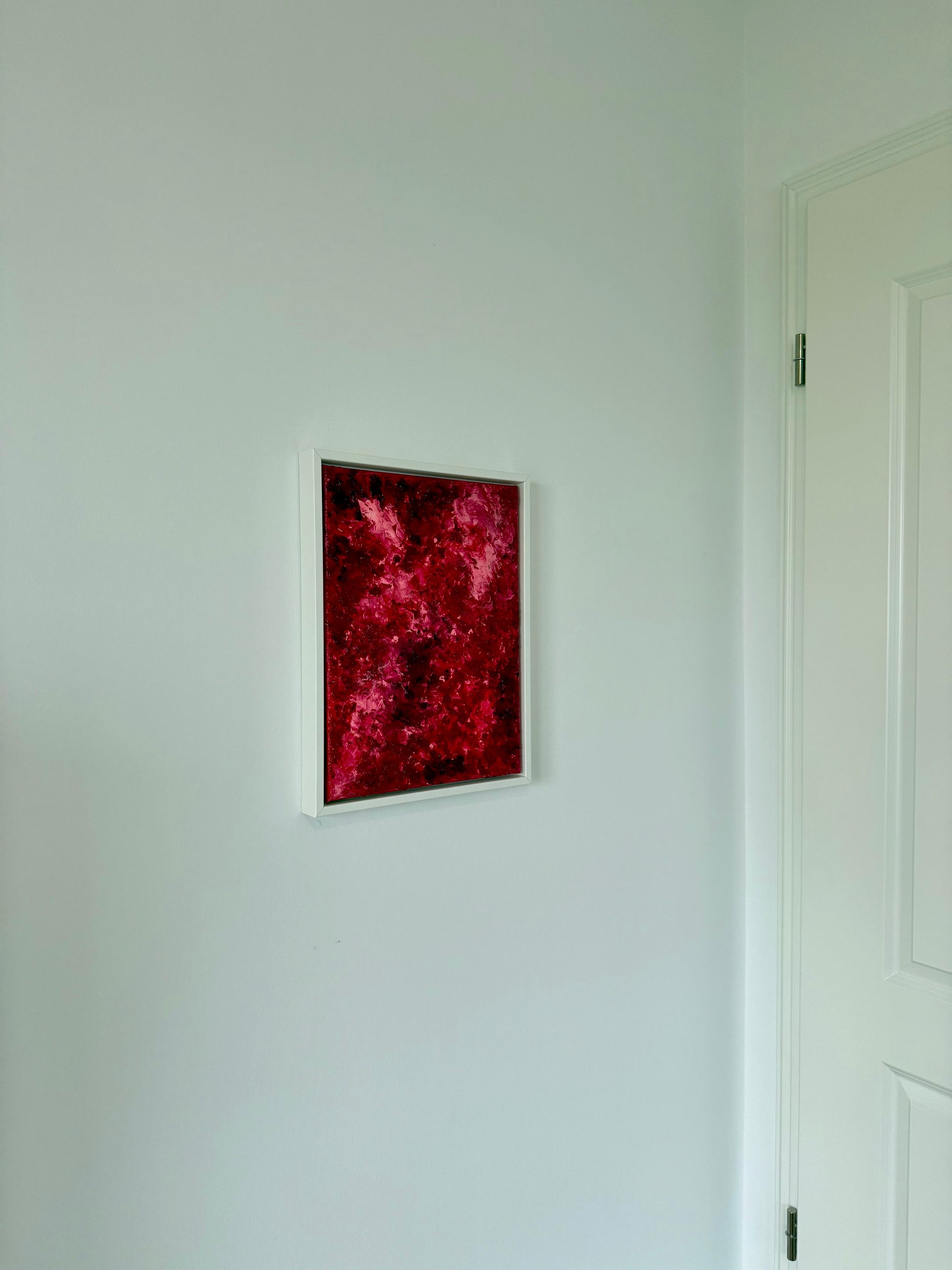 Kleines abstraktes Kunstwerk in kräftigen Rot- und Pinktönen, gerahmt in Weiß, an einer hellen Wand im Wohnraum aufgehängt. Das Bild setzt einen intensiven Farbakzent in einem minimalistischen, ruhigen Interior und wirkt modern sowie klar platziert.