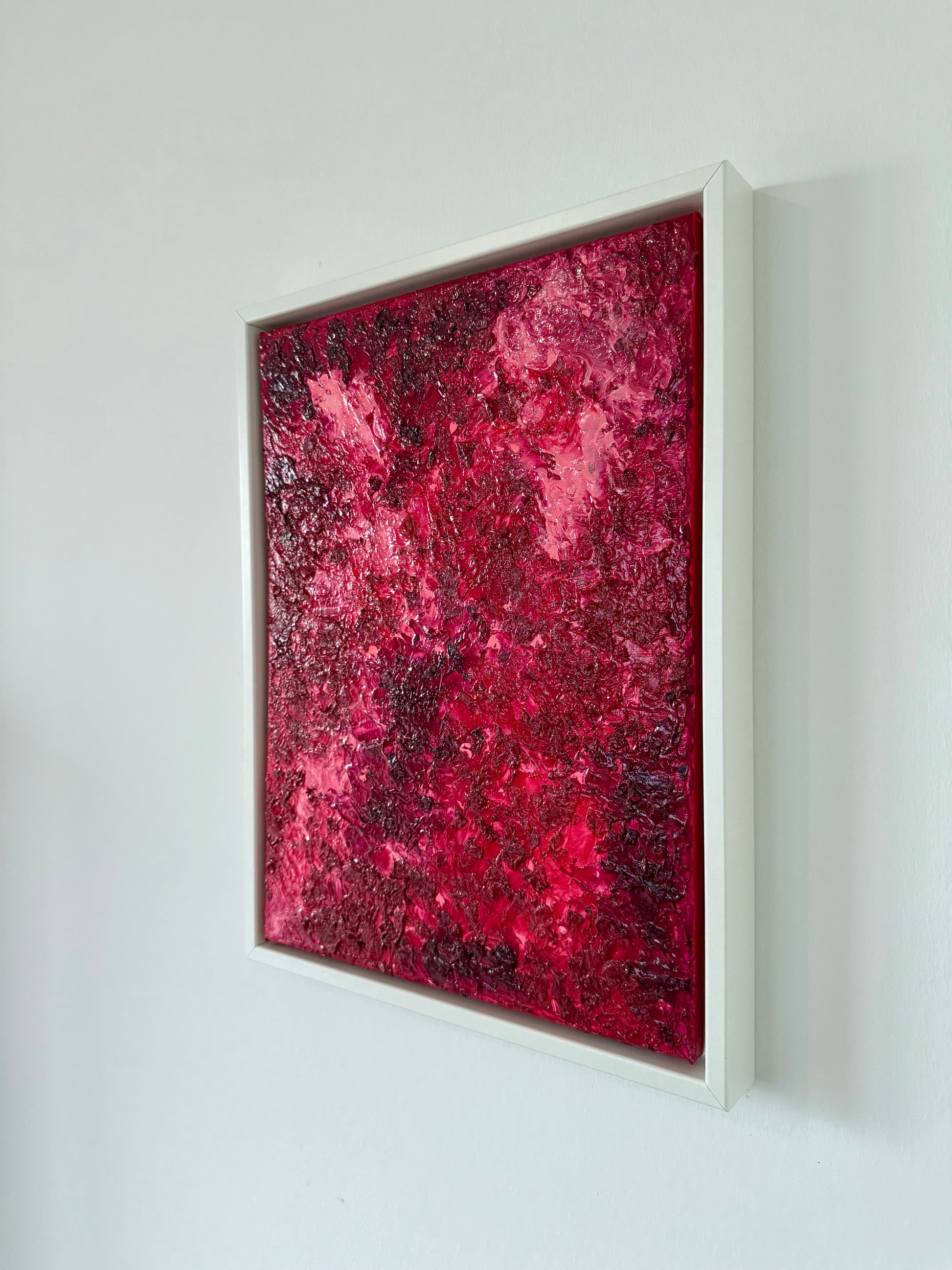 Seitliche Ansicht eines abstrakten Kunstwerks in intensiven Rot- und Pinktönen im weißen Schattenfugenrahmen. Die glänzende, stark texturierte Oberfläche reflektiert das Licht und betont die plastische Tiefe des Werks an der hellen Wand.