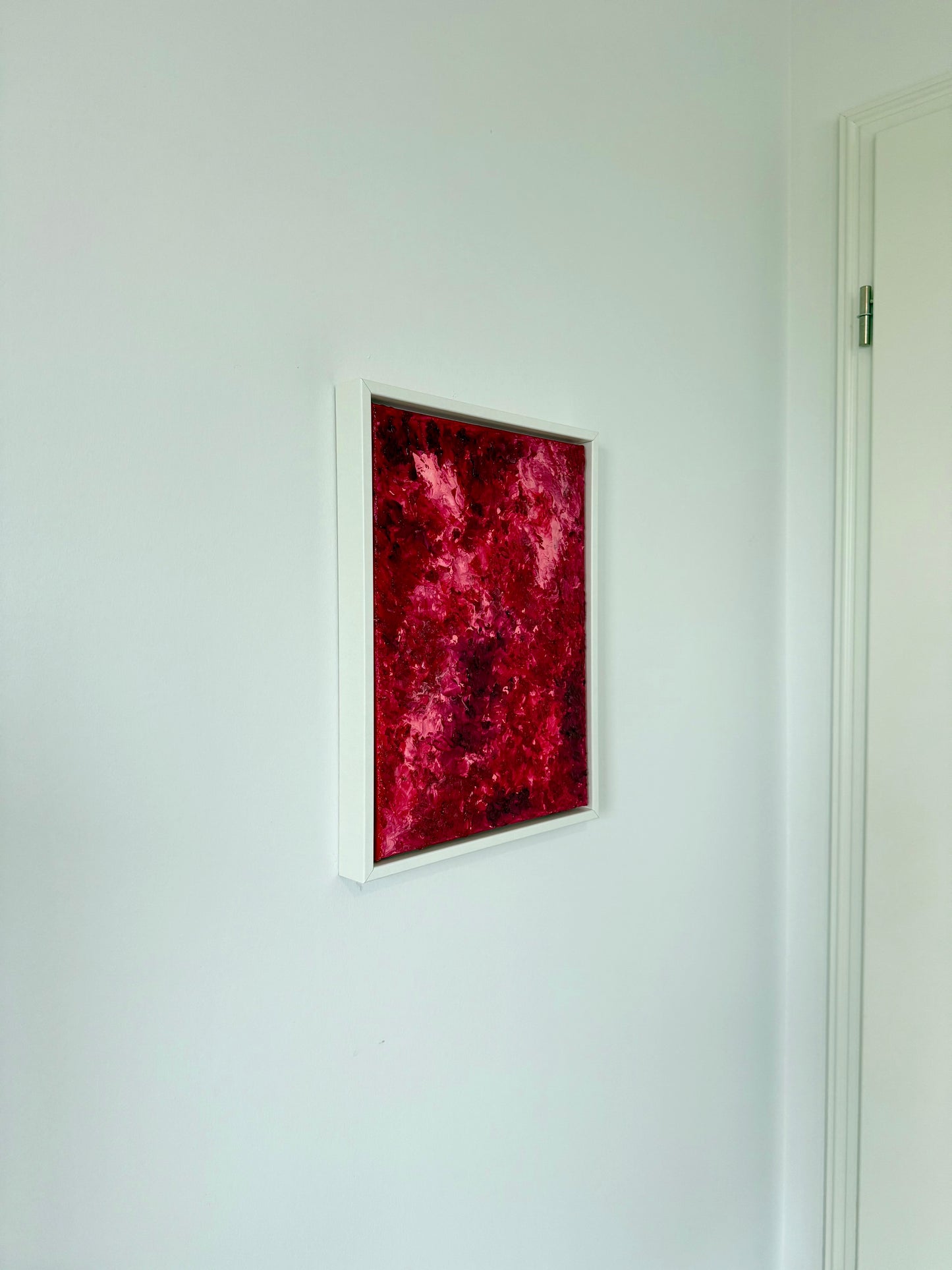 Näher aufgenommene Ansicht eines kleinen abstrakten Kunstwerks in intensiven Rot- und Pinktönen im weißen Rahmen. Das Bild hängt an einer hellen Wand und entfaltet seine Farbwirkung als moderner Akzent in einem reduzierten Wohnambiente.